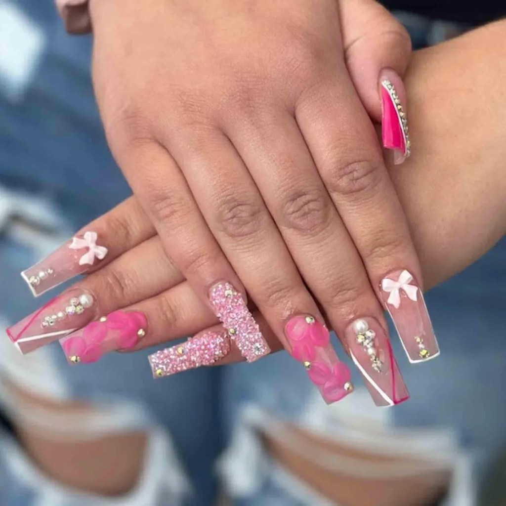 Close-up of hands with elaborate nail art featuring long, embellished pink and clear nails adorned with flowers, rhinestones, and bows.