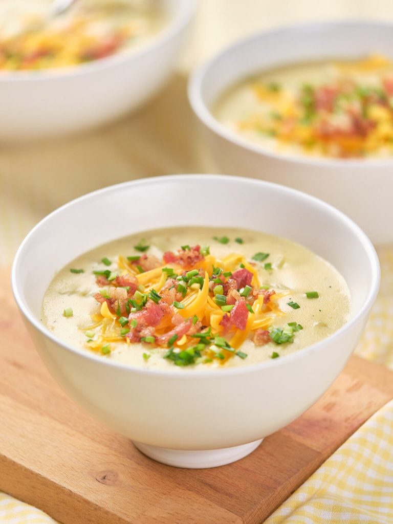 A creamy loaded baked potato soup topped with cheddar cheese, crispy bacon bits, and fresh chives, served in a white bowl against a soft yellow background.
