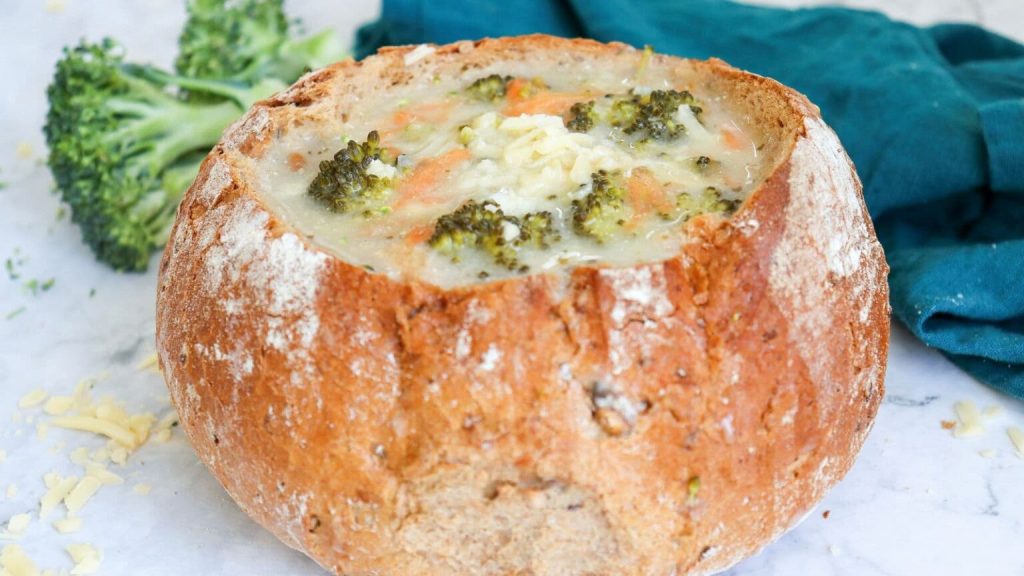 A bowl of creamy broccoli cheddar soup served in a rustic bread bowl, garnished with melted cheese and surrounded by fresh broccoli and a teal napkin.