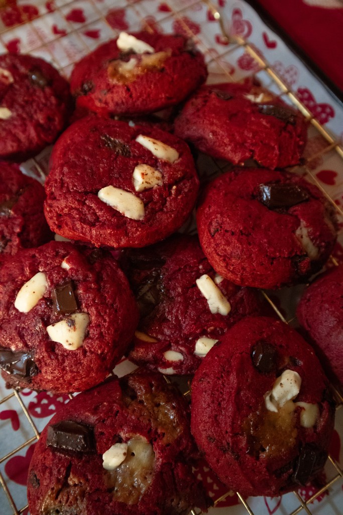 A close-up view of freshly baked red velvet cookies with chunks of dark and white chocolate on a cooling rack.