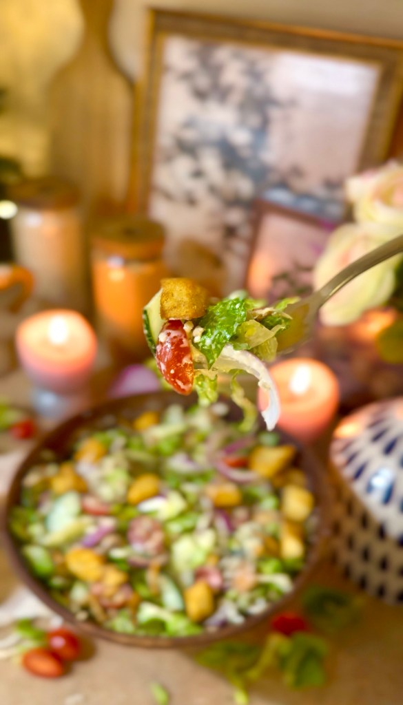 A close-up of a fork holding a bite-sized serving of salad, featuring lettuce, cherry tomatoes, and croutons, with a bowl of salad in the background and candles illuminated for ambiance.