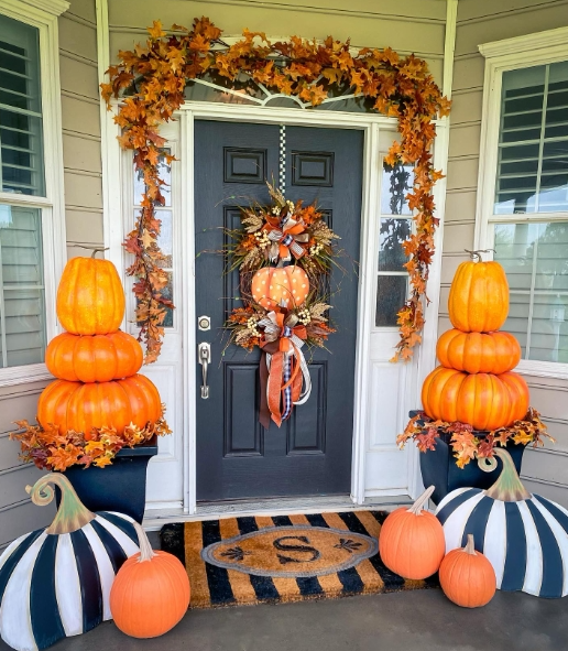 A decorated porch with a dark front door framed by an autumnal wreath and orange leaves, featuring stacked pumpkins in colorful planters and a decorative welcome mat.