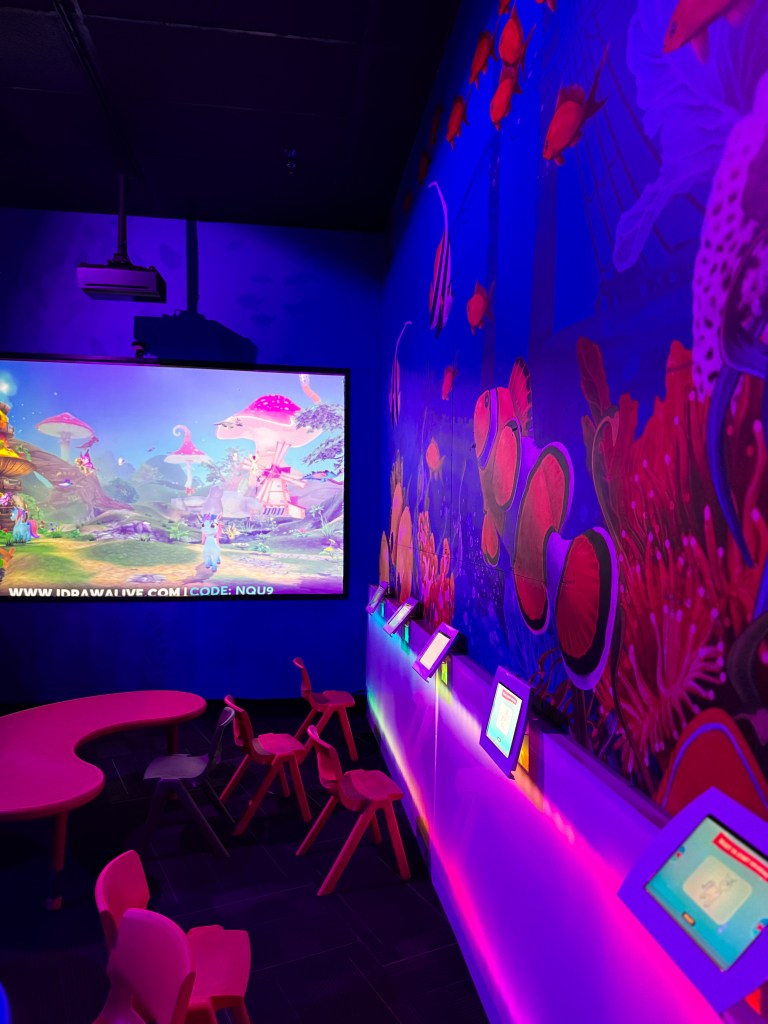 A colorful interactive learning space featuring a large screen displaying animated underwater scenes, vibrant wall murals of marine life, and colorful seating arrangements.