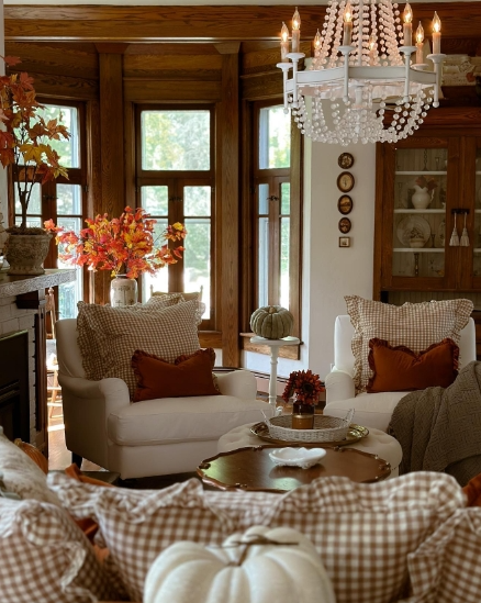 A cozy living room featuring comfortable seating with plaid pillows, warm autumn accents, and a decorative chandelier. The room is enhanced by colorful fall foliage and pumpkins, creating an inviting atmosphere.