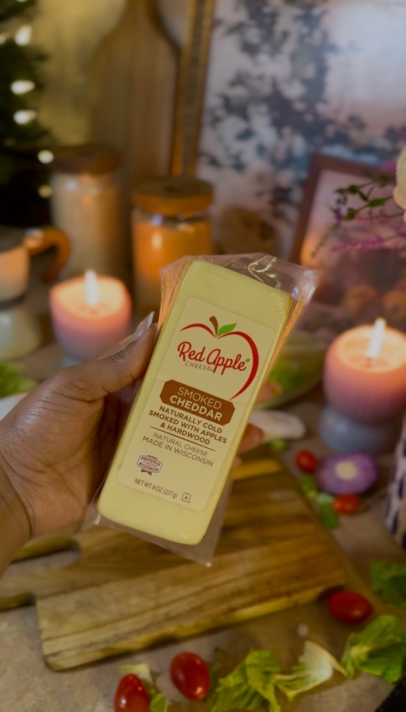 A hand holding a package of Red Apple Smoked Cheddar cheese, surrounded by candles, fresh vegetables, and a cutting board in a warm, cozy setting.