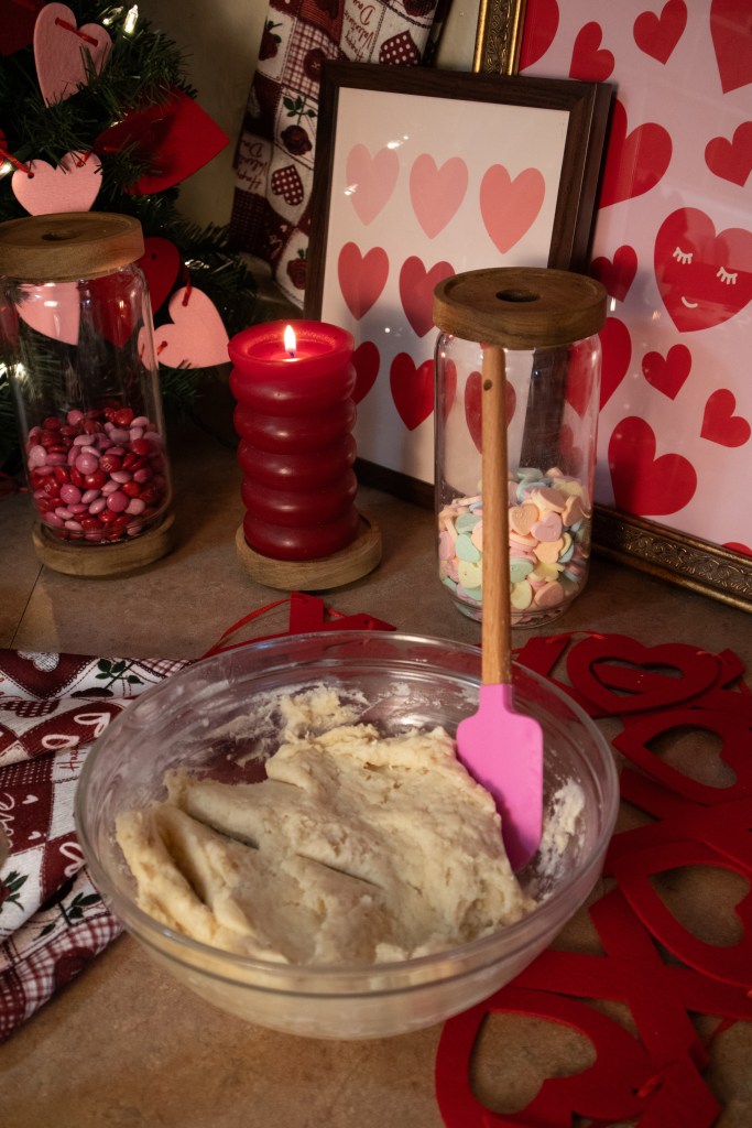 A clear bowl filled with cookie dough, accompanied by a pink spatula, surrounded by decorative elements like red candles, jars of candy hearts, and heart-themed decor.