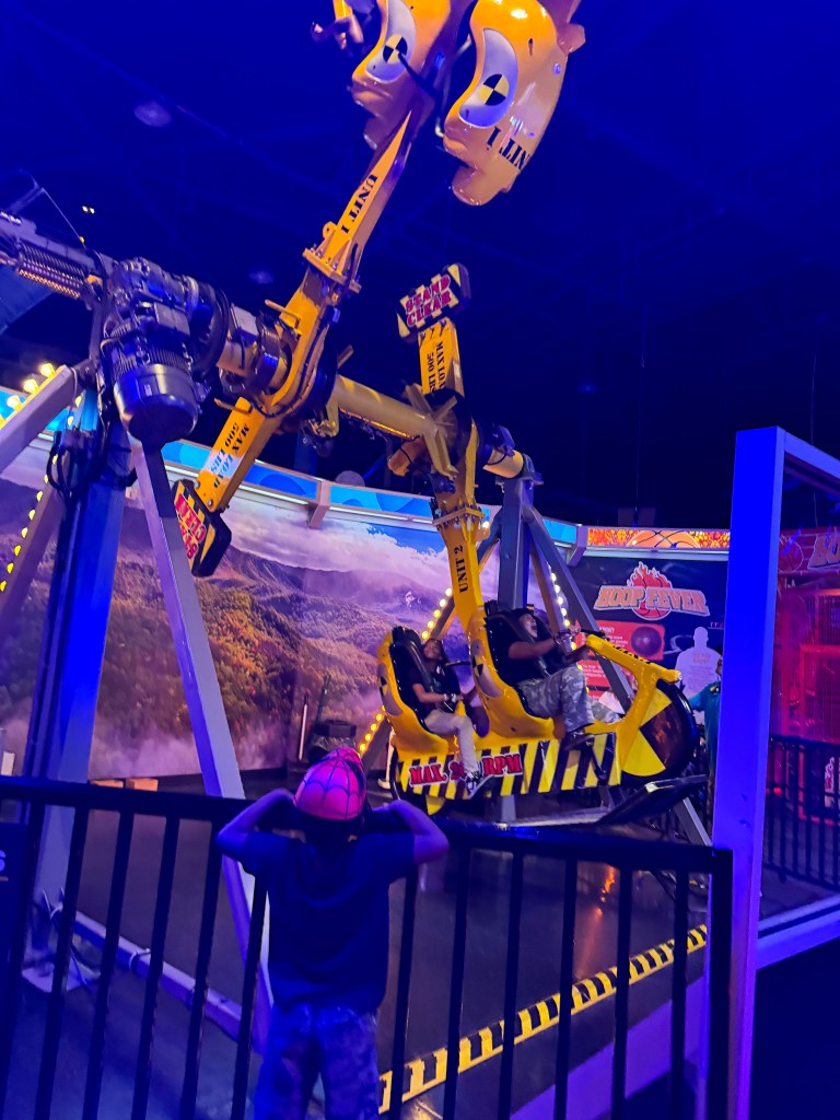 A child wearing a spider-themed hat watches excitedly as riders enjoy a thrilling amusement ride featuring robotic arms and yellow seats, set against a colorful backdrop in a dimly lit arcade.