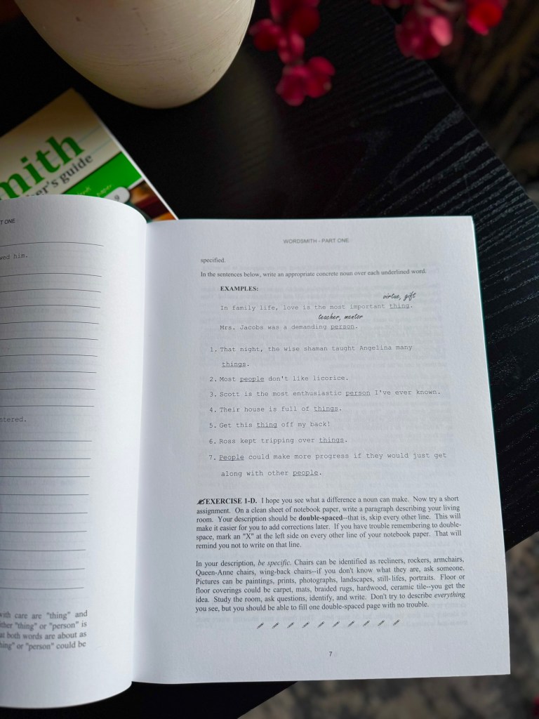 A close-up view of a workbook page titled 'Wordsmith - Part One,' featuring examples and exercises about identifying concrete nouns. A potted plant and a book are visible in the background.