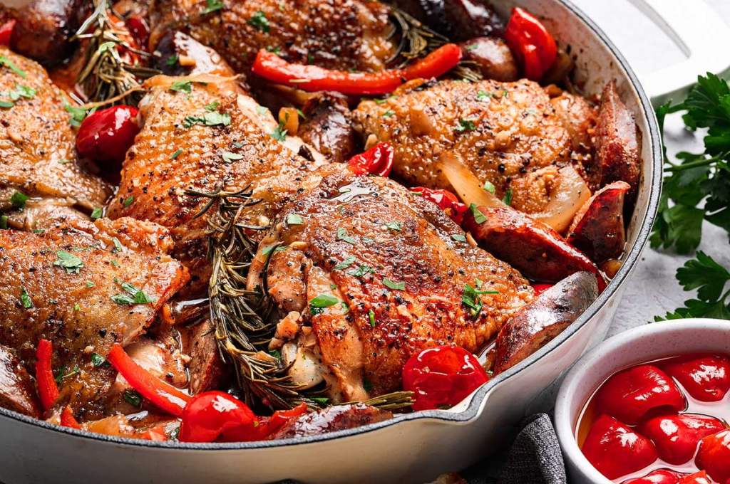 A close-up view of a dish featuring golden-brown chicken thighs, garnished with fresh herbs and surrounded by colorful bell peppers and cherry tomatoes in a rustic pot.