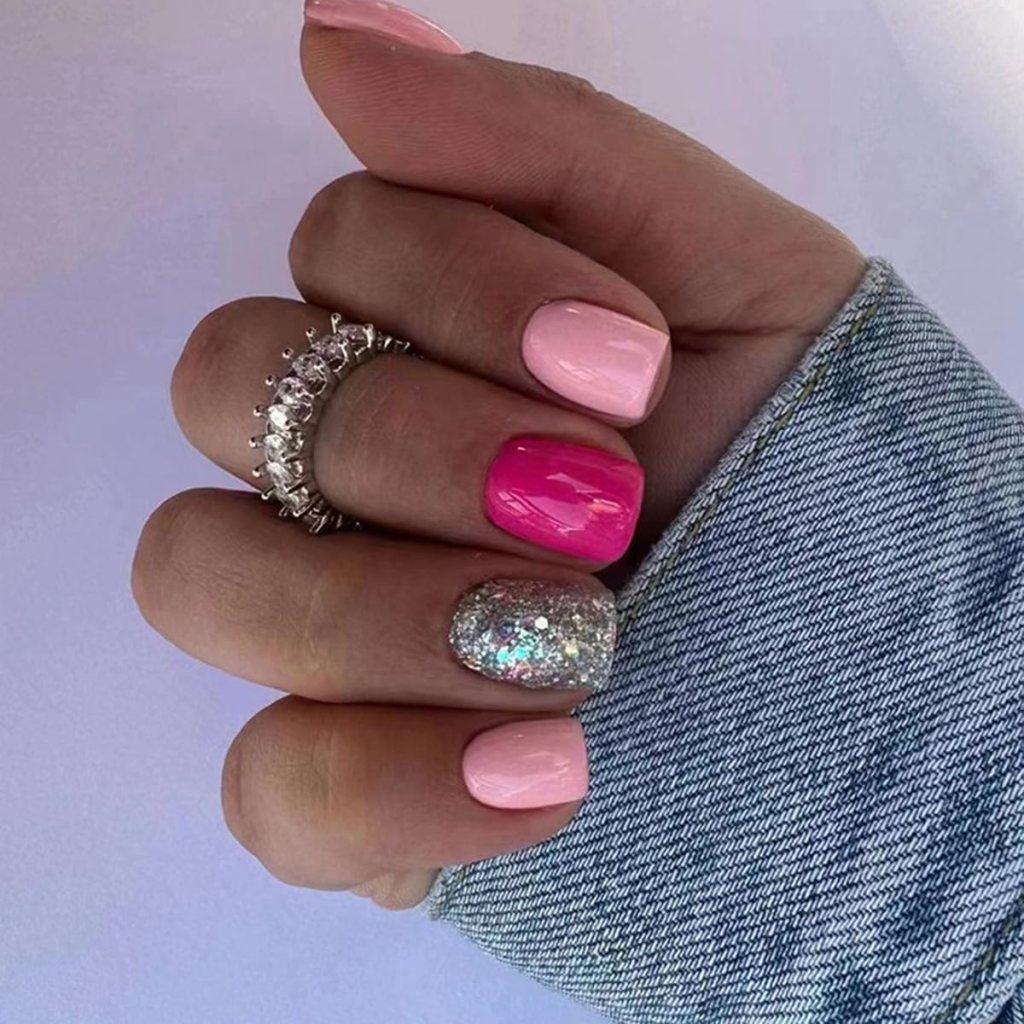 Close-up of a hand displaying manicured nails in various shades of pink and a glittery silver nail, wearing a decorative ring, with a denim sleeve visible in the background.