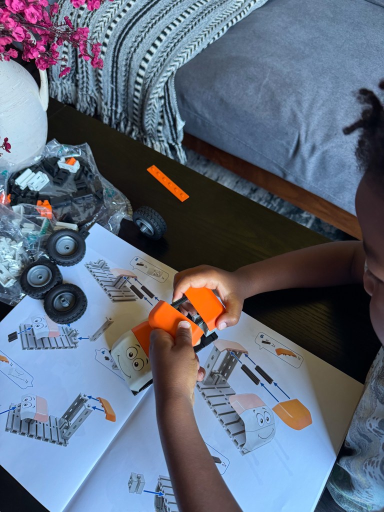 A child is assembling a toy using colorful blocks while following instruction images in a booklet, surrounded by additional toy pieces and a decorative vase.