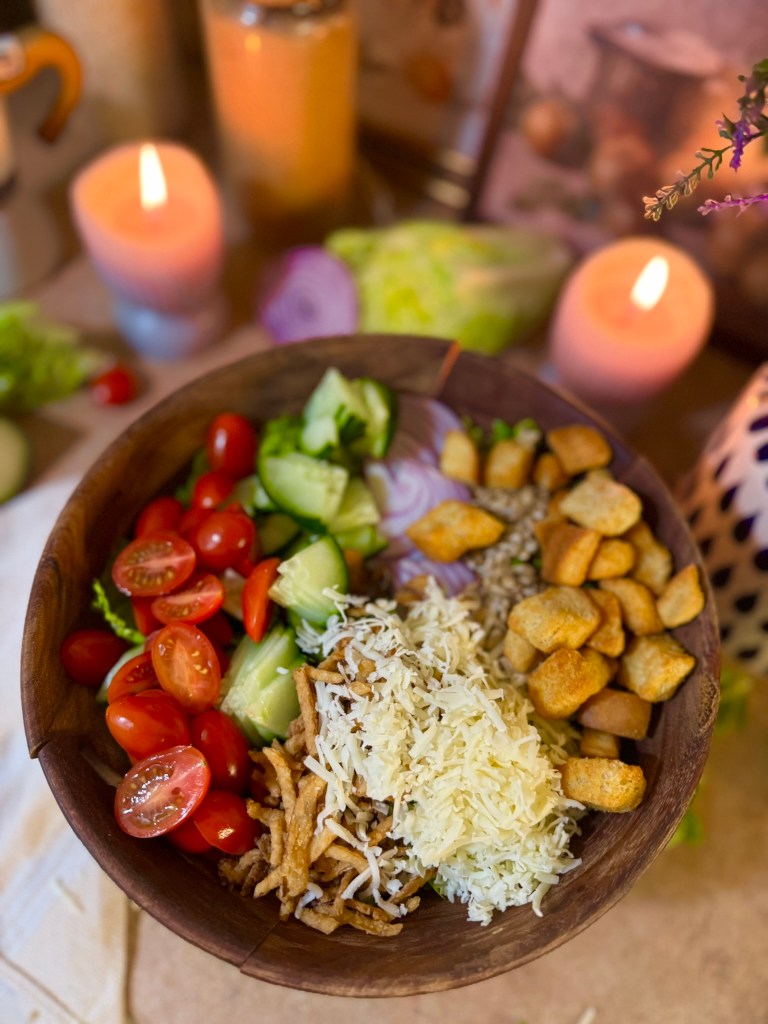 A wooden bowl filled with a colorful salad featuring cherry tomatoes, sliced cucumbers, grated cheese, fried onions, croutons, and sunflower seeds, with candles and fresh vegetables in the background.
