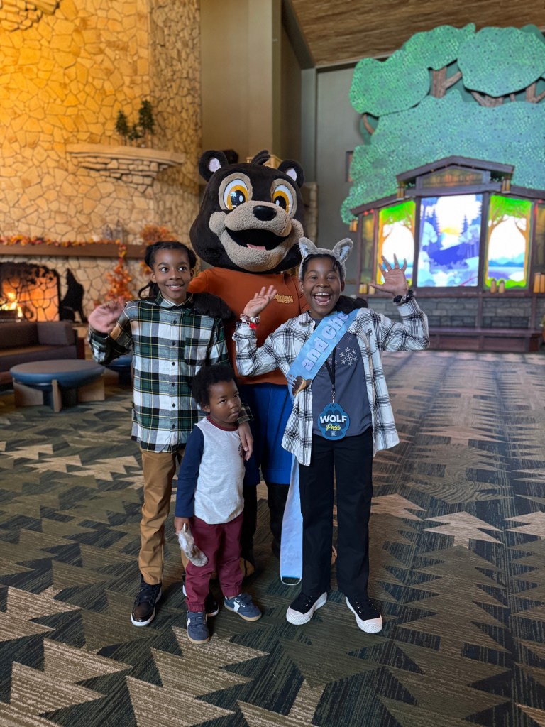 A family of three children posing happily with a friendly bear mascot at Great Wolf Lodge, with a warm lobby atmosphere featuring a stone fireplace and a colorful backdrop.