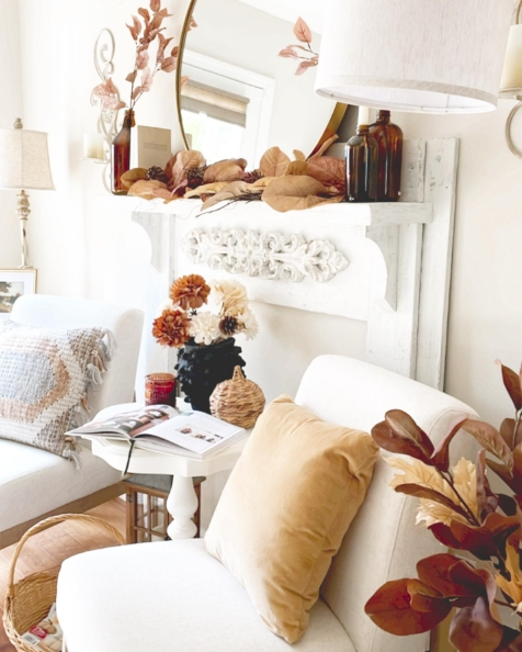 Cozy living room corner featuring a white chair with a mustard-colored pillow, a small table with a magazine, and autumn-themed decor on a mantel, including dried leaves and flowers.