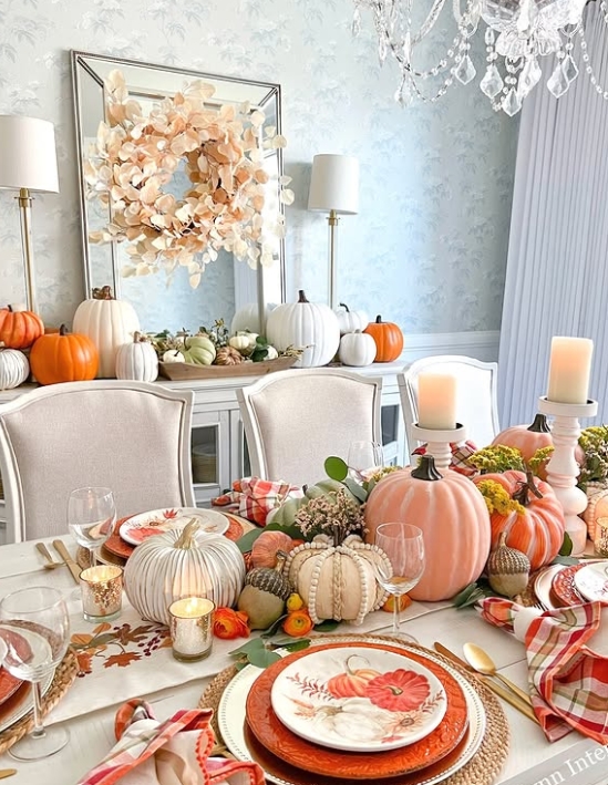 A beautifully styled Thanksgiving table featuring a variety of pumpkins, elegant dinnerware, and a floral centerpiece. The setting includes warm tones, candles, and a mirror reflecting a festive atmosphere.
