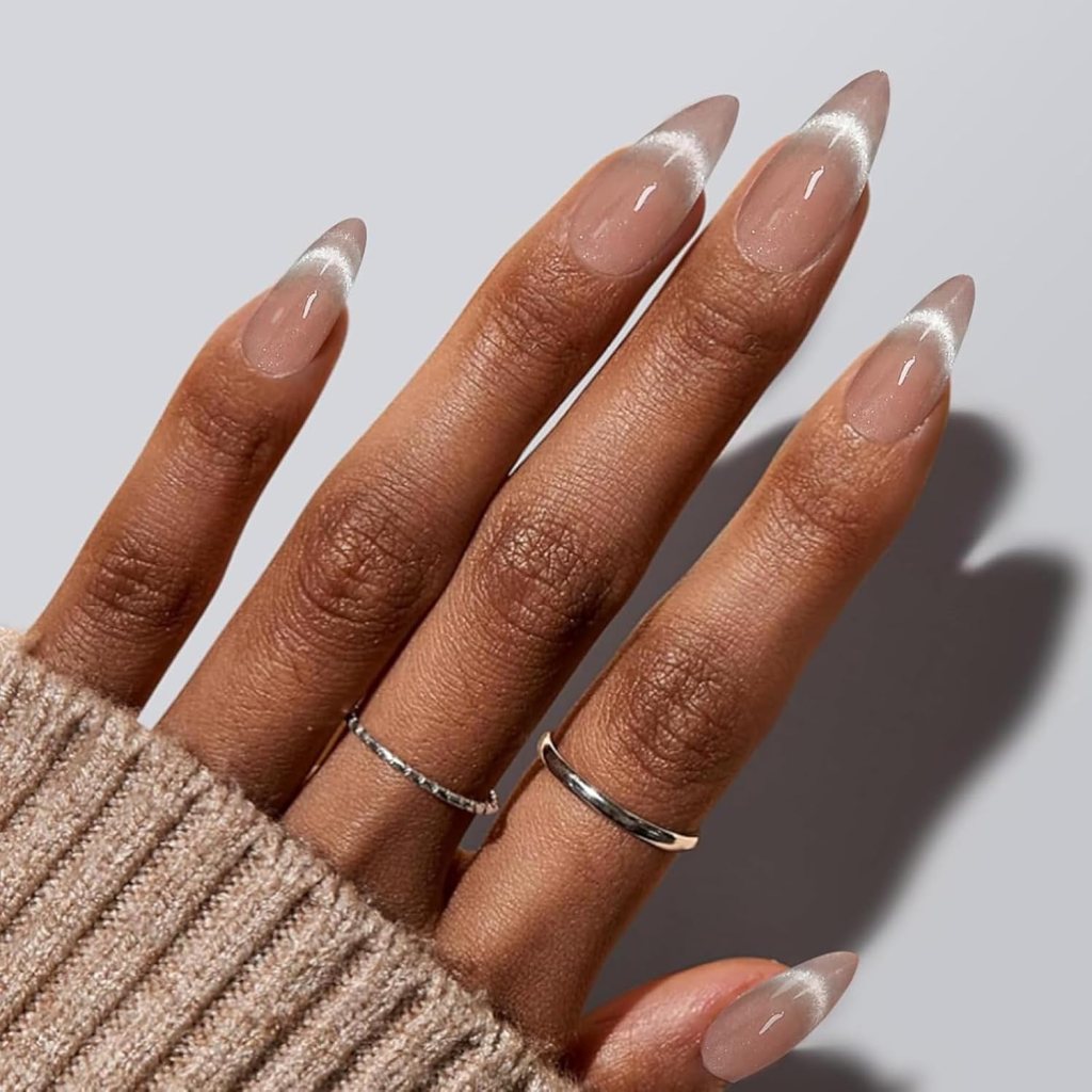 Close-up of a hand with almond-shaped nails painted in a nude color with white tips, wearing two silver rings, and partially covered by a beige knitted sweater.