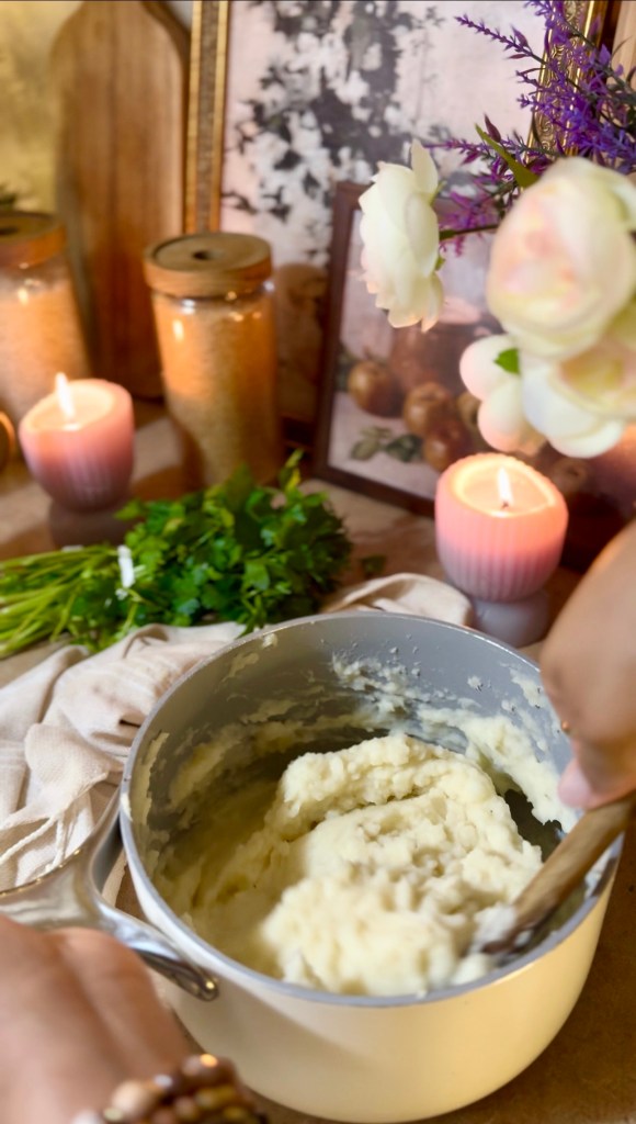 A person stirring creamy mashed potatoes in a pot, surrounded by candles, herbs, and decorative items.