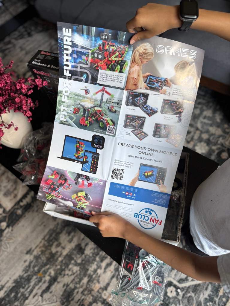 A child holding a brochure for the Fischer Technik STEM Kit, showcasing its features and design options, with a box of materials and various building pieces visible in the background.