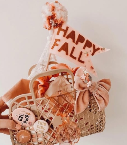 A pastel basket filled with seasonal accessories including hair ties, pins, and a flag that says 'HAPPY FALL'.