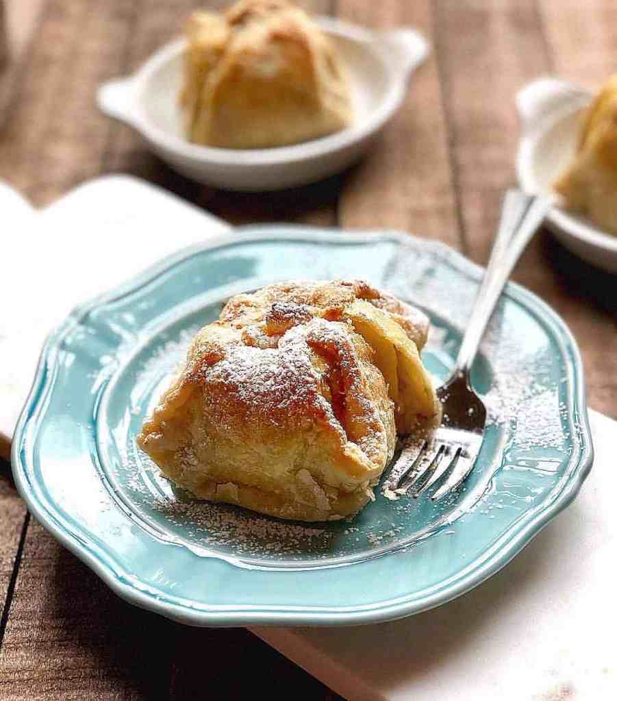 A warm apple dumpling dusted with powdered sugar on a blue plate, with a fork nearby, set on a wooden table.