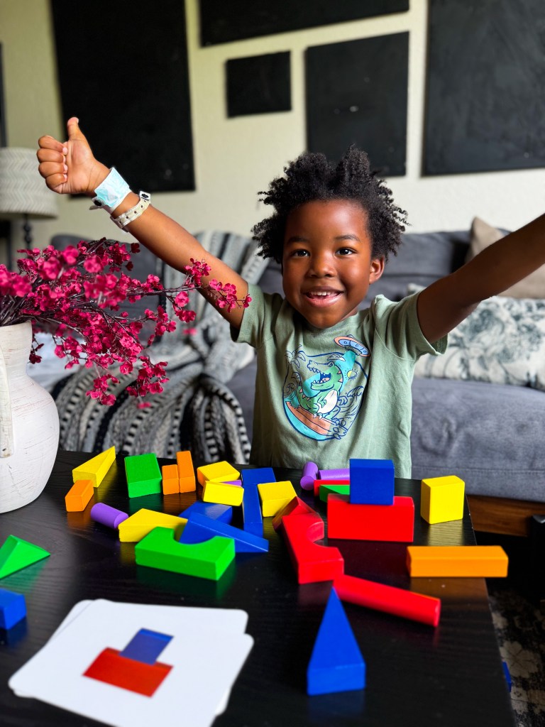 A joyful child with curly hair raises their arms in excitement while sitting at a table covered with colorful wooden building blocks. A vase with pink flowers is in the foreground, and shapeshifting cards can be seen on the table.