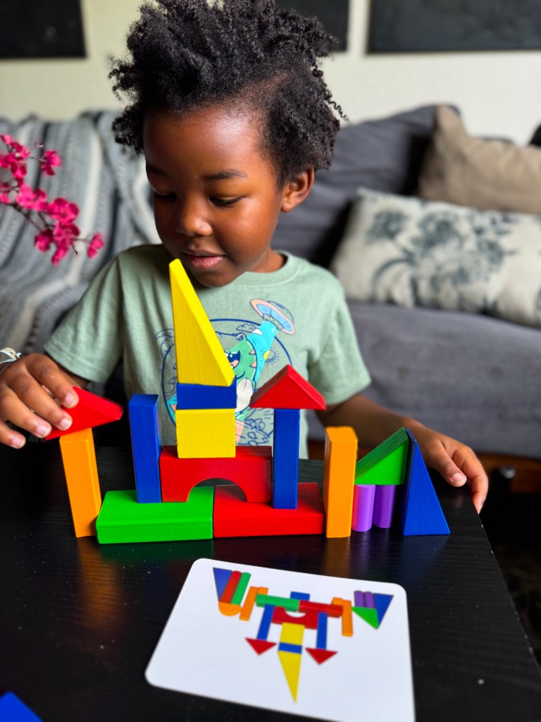 A young child focused on building a colorful structure with various shapes of blocks, seated at a table with a guidance card showing a similar design.