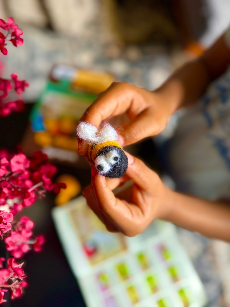 A child's hand holding a handmade needle-felted bee figure, surrounded by colorful wool fibers and a booklet in the background, with pink flowers adding a vibrant touch.