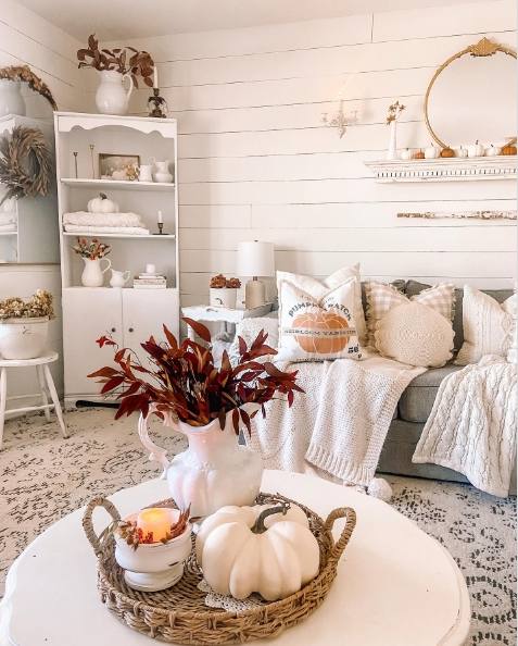 A cozy living room featuring a light-colored couch adorned with textured pillows, a soft throw, and a decorative arrangement of autumn foliage in a white vase on a coffee table with pumpkins and a candle.