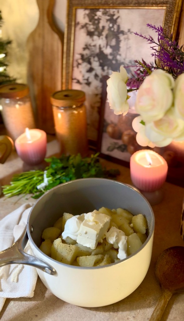 A pot filled with diced potatoes topped with sour cream and cubes of butter, surrounded by candles, herbs, and decorative elements.