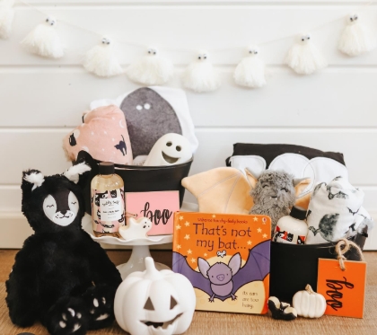A collection of Halloween-themed goodies arranged in a cozy setting. Features include plush toys, a children’s book titled 'That's Not My Bat,' a small white pumpkin, and decorative elements with a black and white color scheme, all placed near a garland of ghost figurines.