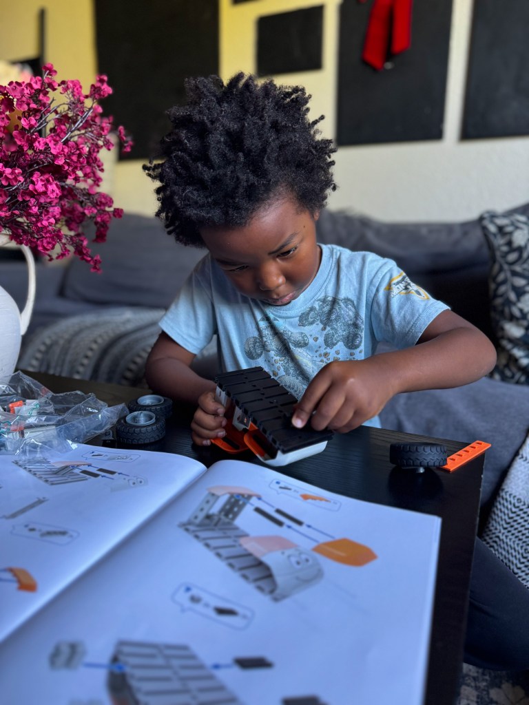 A young child with curly hair is focused on assembling a toy car using building instructions, surrounded by parts and tools on a table.
