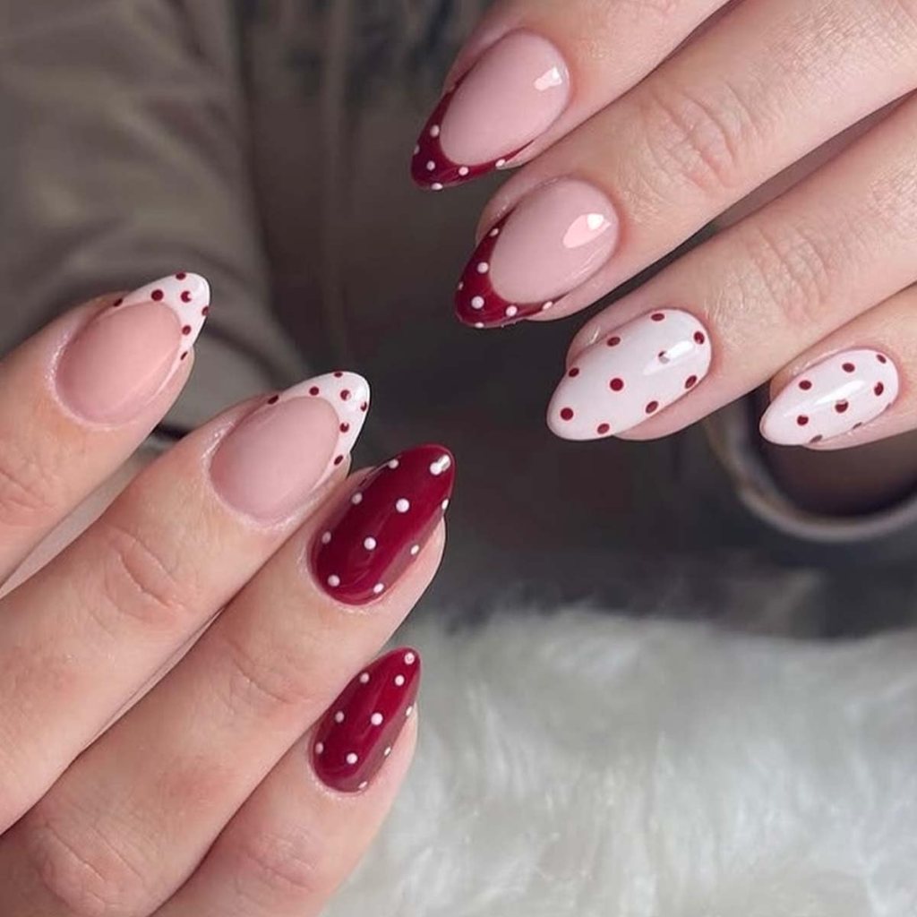A close-up image of two hands showing stylish manicured nails. The nails feature a mix of nude, red, and white colors with various designs including pointed tips, polka dots, and half-moon patterns.