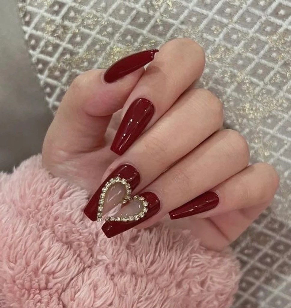 A hand with long, glossy red nails featuring a heart-shaped design on one nail, adorned with rhinestones, resting on a soft pink fabric.