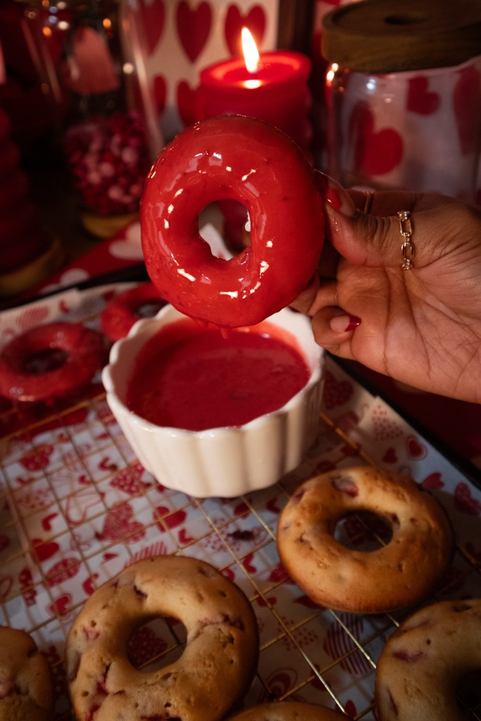 A hand holding a red frosted donut above a bowl of pink dipping sauce, with more donuts on a wire rack in the background.