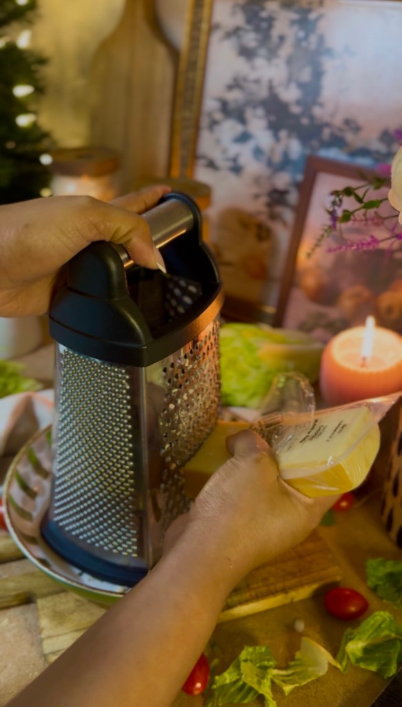 A person's hand holding a package of cheese while grating it using a metal cheese grater, with fresh ingredients like lettuce and cherry tomatoes in the foreground.