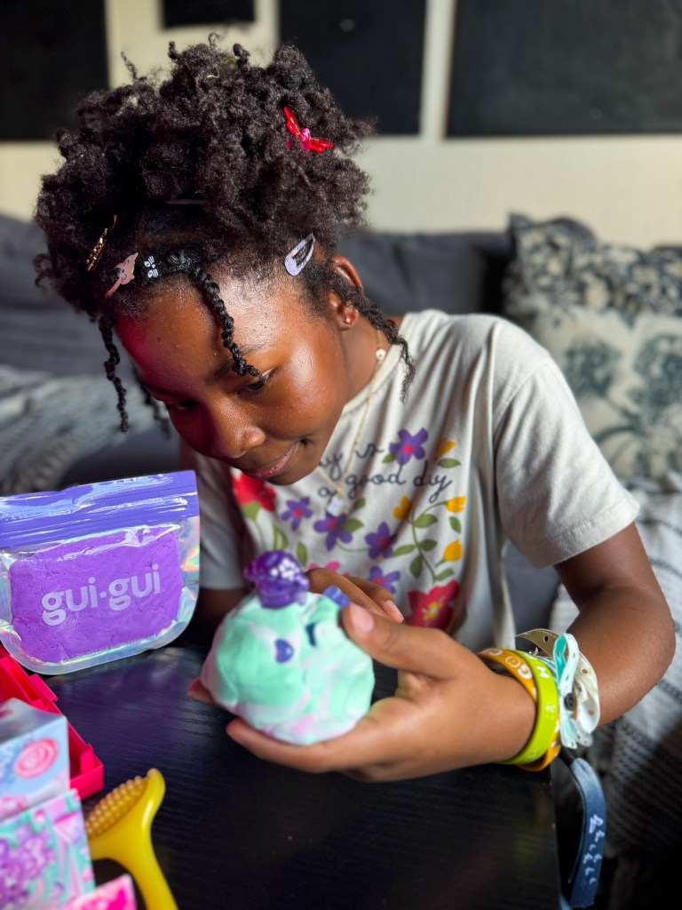 A young girl with curly hair styled with colorful clips observes a multicolored dough figure she holds in her hands, surrounded by crafting materials on a table.