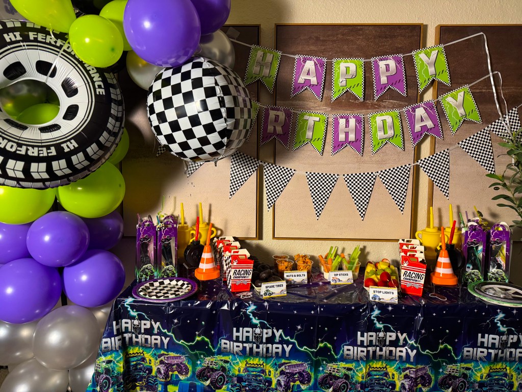 A birthday party setup featuring a colorful backdrop with a tire-themed balloon arrangement, a 'Happy Birthday' banner, and a decorated table with racing-themed snacks and decorations.