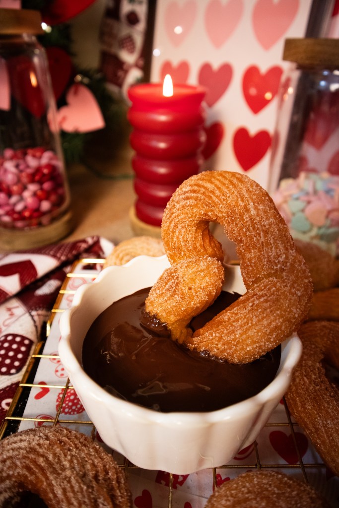 A churro dipping into a bowl of chocolate sauce, with a red candle and heart-themed decorations in the background.