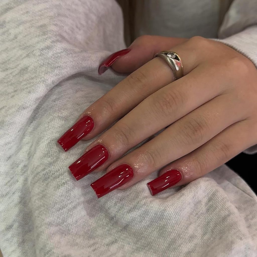 A close-up of a hand with long, glossy red nails, adorned with a silver ring, resting on a gray sweater.