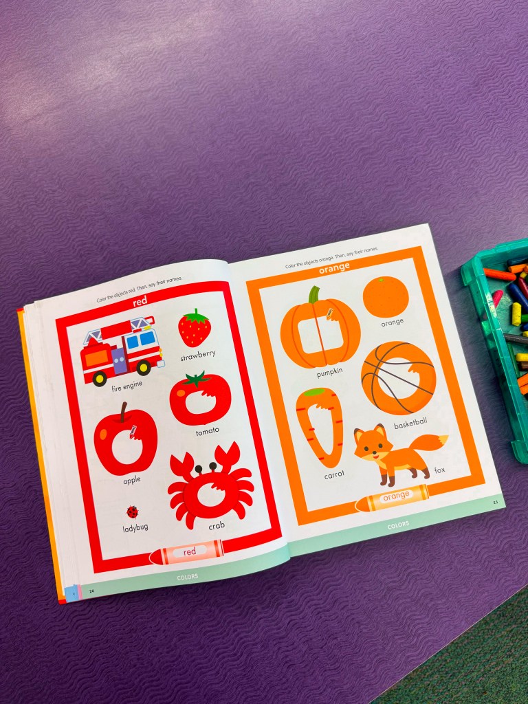Open book page displaying illustrations of red and orange objects for coloring, including a fire engine, strawberries, apples, pumpkins, and a fox. Crayons are visible in a container nearby.