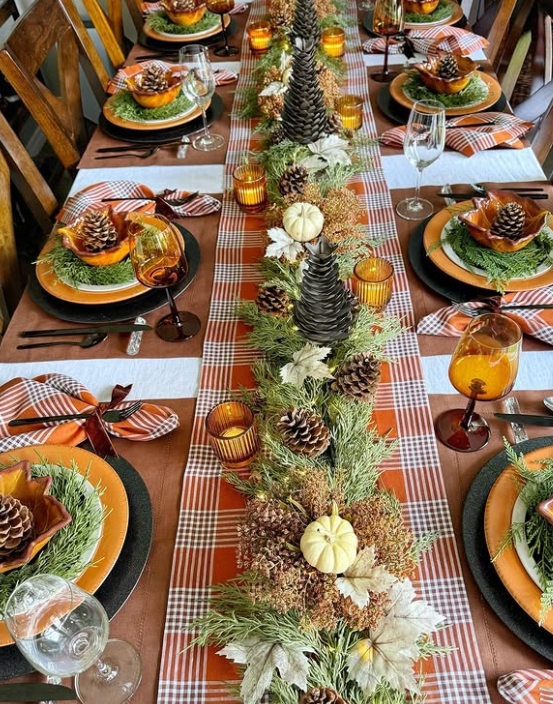 An elegant Thanksgiving tablescape featuring a plaid table runner adorned with pinecones, greenery, and miniature pumpkins. The table settings include orange plates, amber glassware, and checked napkins, creating a warm and inviting atmosphere.