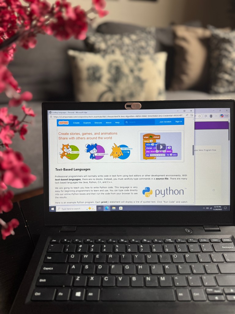 A laptop on a table displays a coding lesson focused on text-based programming languages like Python, with colorful graphics and examples from the Scratch platform in the foreground. A flower arrangement is visible in the left corner, and a soft couch is blurred in the background.