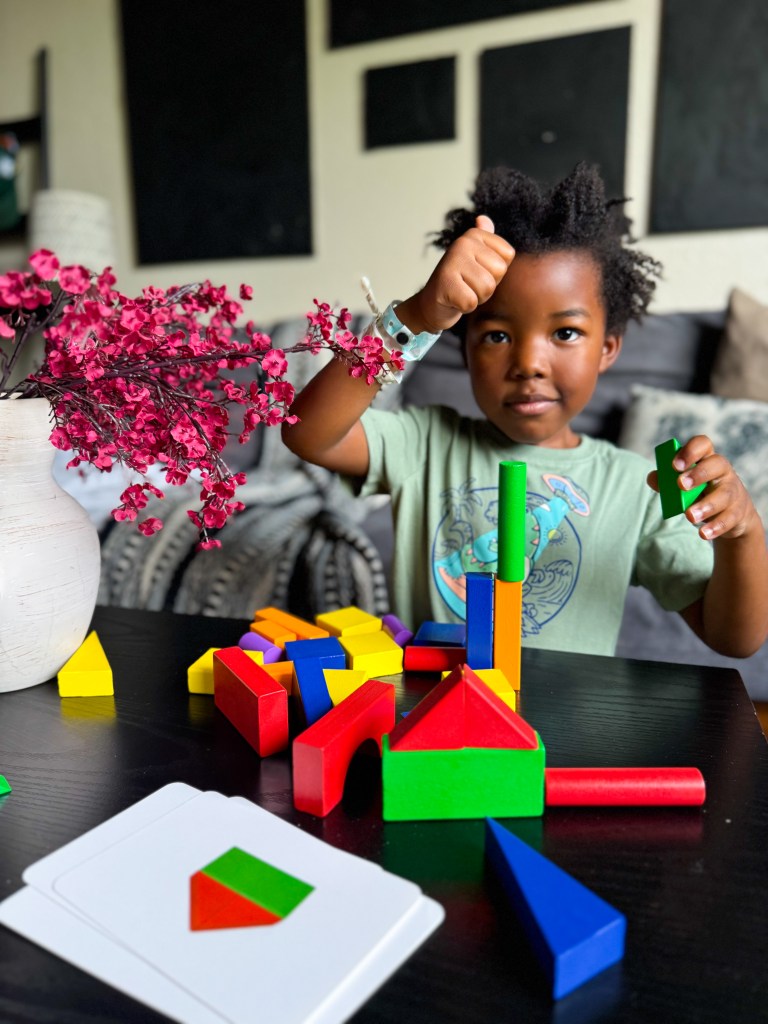 A young child with short curly hair is enthusiastically stacking colorful geometric blocks on a table. A few cards with shapes are visible in front of them, as well as a vase with pink flowers.