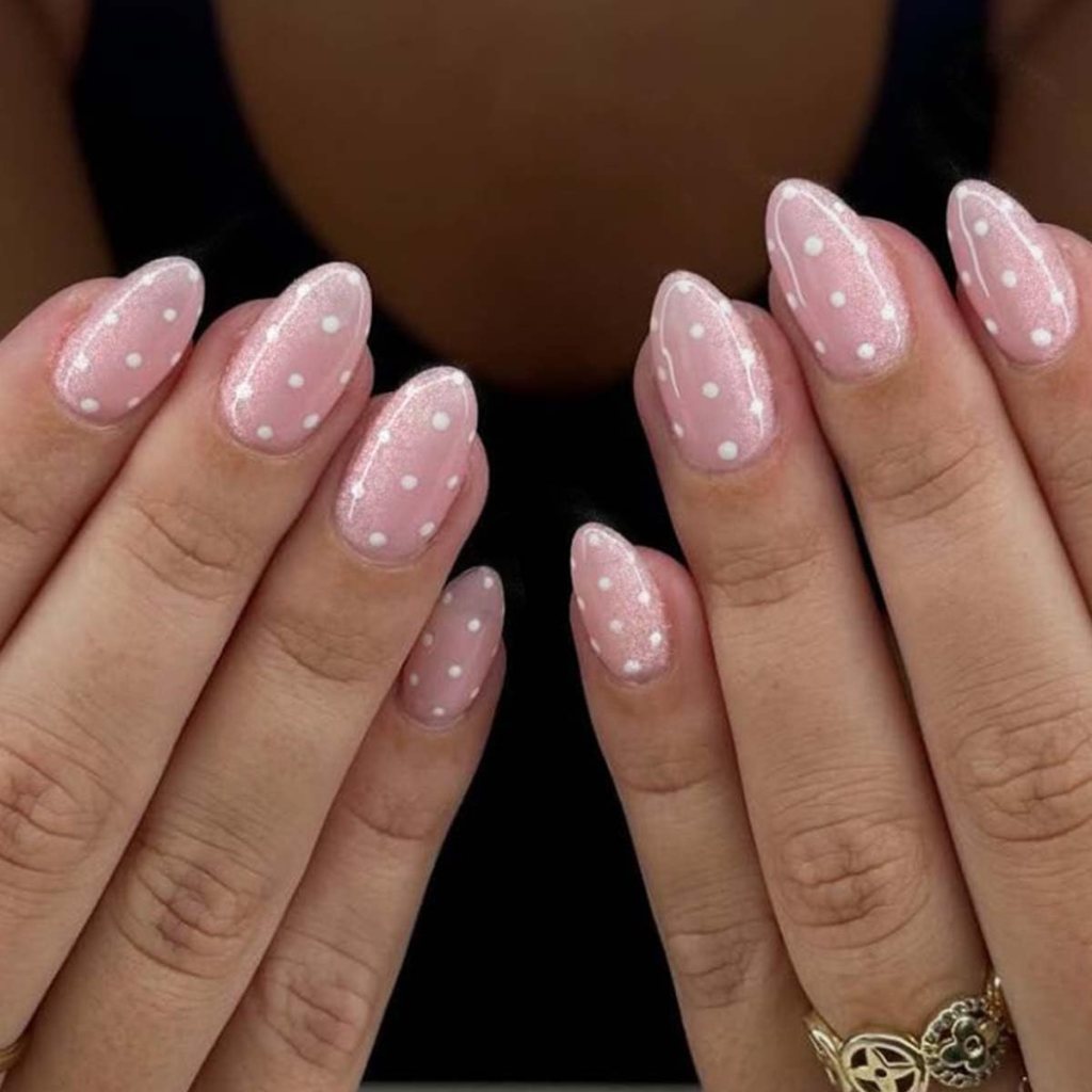 Close-up of two hands with polished almond-shaped nails in a light pink color, featuring white polka dots and a subtle glitter finish.