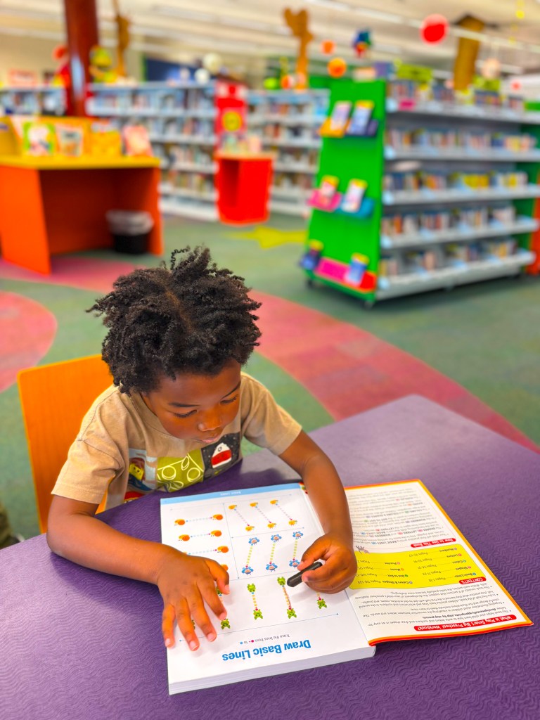 A young child with curly hair sitting at a table in a colorful library, drawing in a workbook focused on basic line drawing activities.