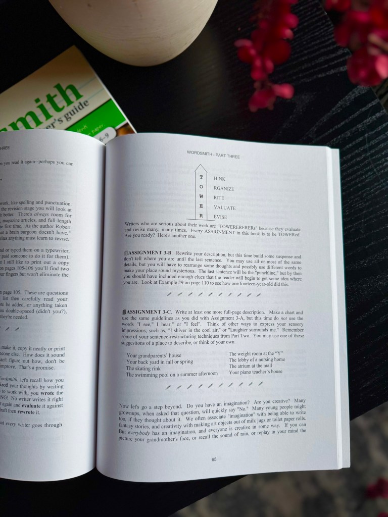 A close-up view of an open book titled 'Wordsmith - Part Three', featuring writing assignments and explanations. A green book partially visible in the background and a decorative vase with flowers are also present.
