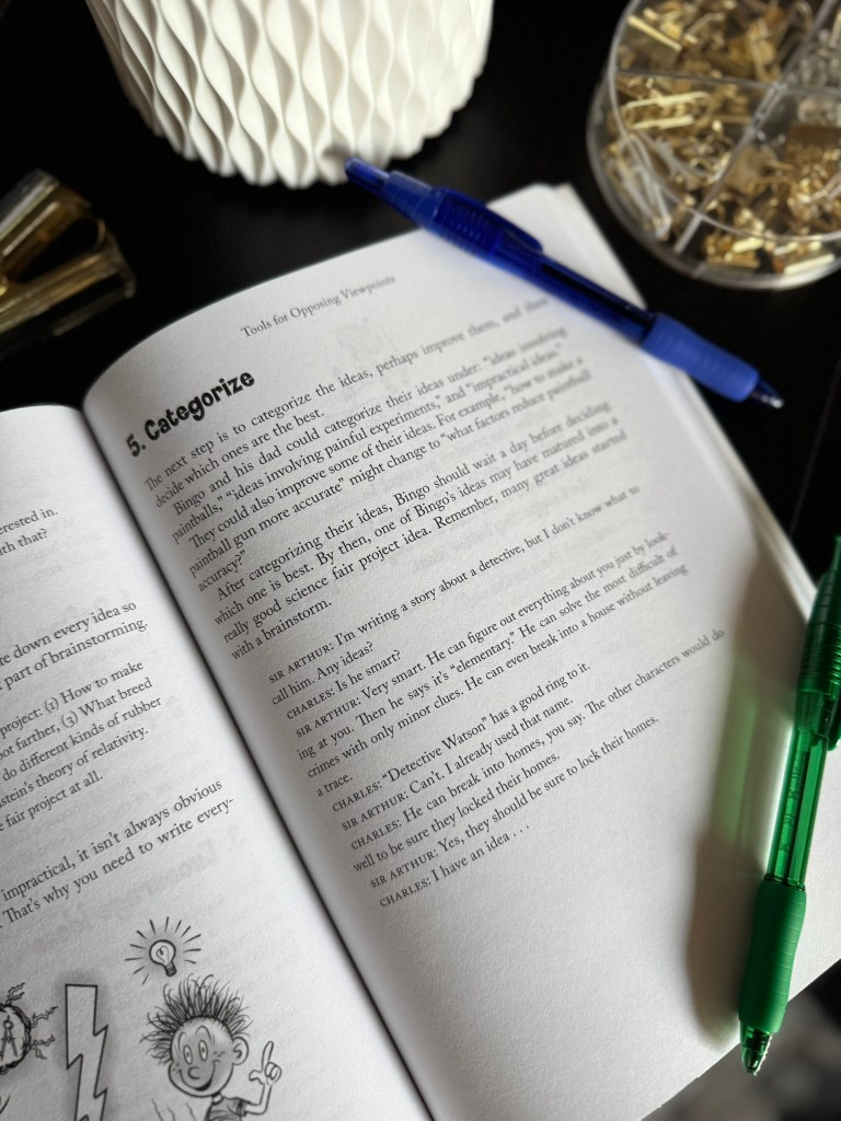 An open book displaying a lesson on categorizing ideas, accompanied by a blue pen and a green pen, with a decorative item and paper clips in the background.