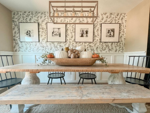 A rustic dining table set with a natural wood finish, featuring a centerpiece of decorative pumpkins and greenery. The backdrop includes framed black-and-white photos and a subtle leaf-patterned wallpaper, complemented by black chairs and a stylish light fixture.
