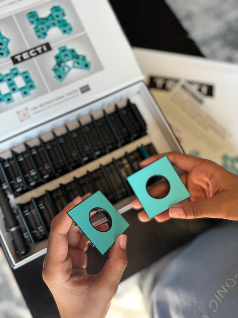 Two turquoise cubes with circular openings being held above an organized tray of similar pieces inside a box labeled 'TECTI'.
