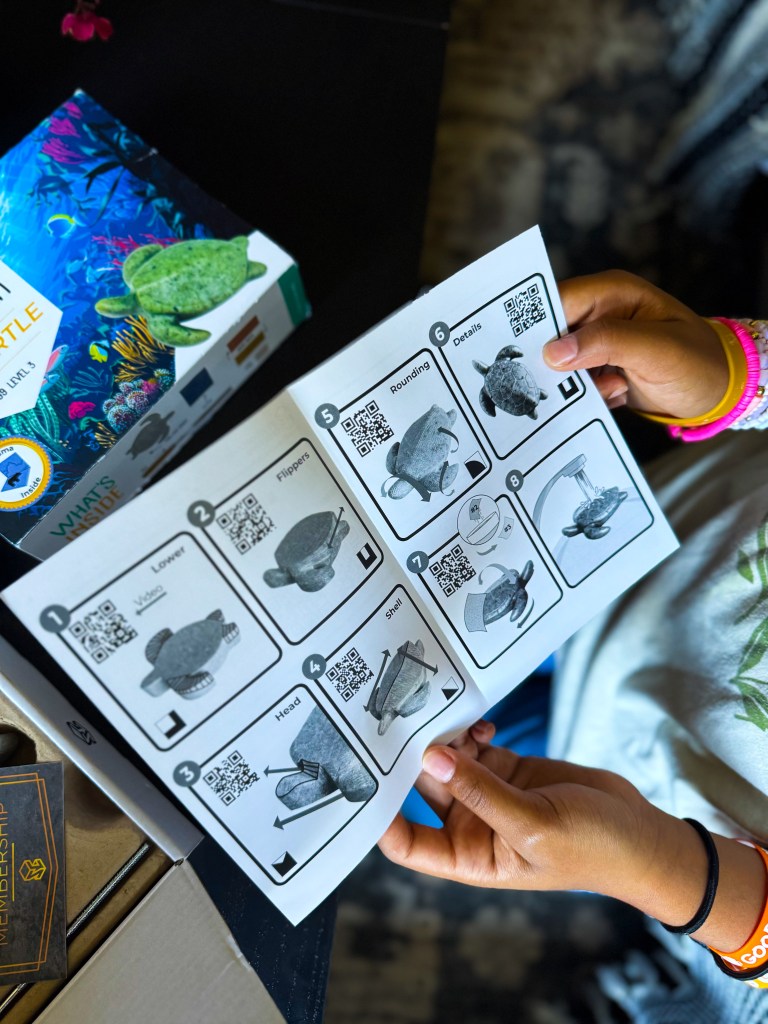 A person holding an instruction manual with steps to assemble a turtle figure, featuring various illustrations and QR codes for guidance. In the background, a colorful turtle-themed box and a blurred hand with a bracelet.